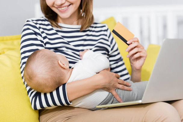 Küçük oğlu olan gülümseyen bir kadının kanepenin üzerinde otururken dizüstü bilgisayarının yanında kredi kartı tutması. - Fotoğraf, Görsel