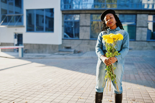 Stylish fashionable african american women in jeans wear and black beret with yellow flowers bouquet posed outdoor in sunny day against blue modern building. - Fotografie, Obrázek