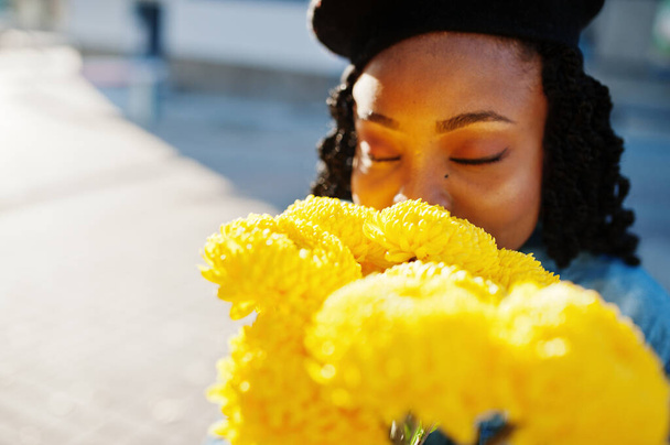 Stylish fashionable african american women in jeans wear and black beret with yellow flowers bouquet posed outdoor in sunny day against blue modern building. - Foto, Imagen