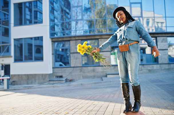 Stylish fashionable african american women in jeans wear and black beret with yellow flowers bouquet posed outdoor in sunny day against blue modern building. - Foto, immagini