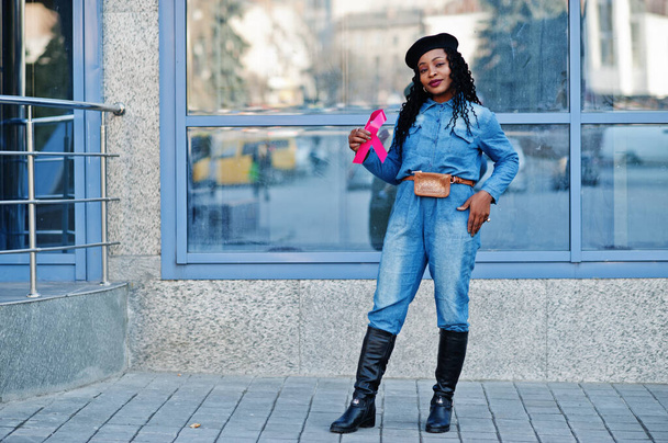 Síla bojovat. Stylová módní africká americká žena v džínách a černý baret proti moderní budově s růžovou stuhou na rakovinu prsu. - Fotografie, Obrázek