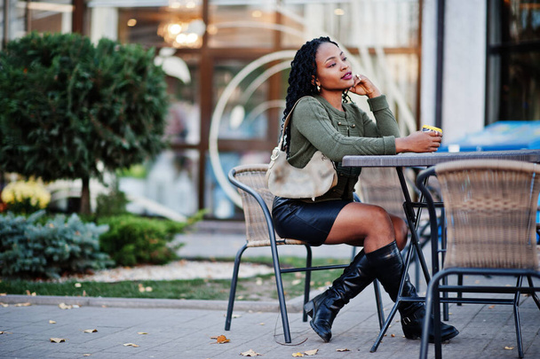 Stylish fashionable african american women in green sweater and black skirt posed outdoor cafe, sitting by table with cup of coffee. - Foto, imagen