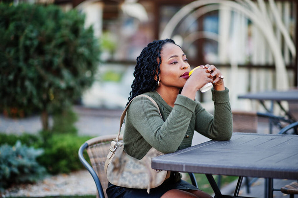 Stylish fashionable african american women in green sweater and black skirt posed outdoor cafe, sitting by table with cup of coffee. - Foto, Imagem