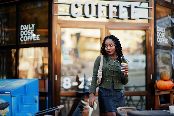 Stylish fashionable african american women in green sweater and black skirt posed outdoor cafe with cup of coffee and speak on mobile phone. - Foto, immagini