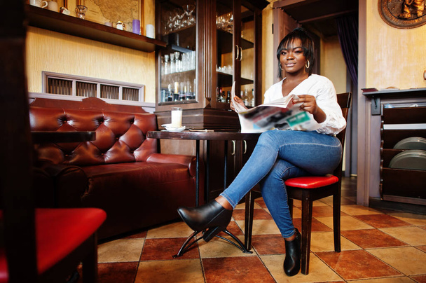 Stylish african american women in white blouse and blue jeans posed at cafe with newspaper. - Foto, Imagem
