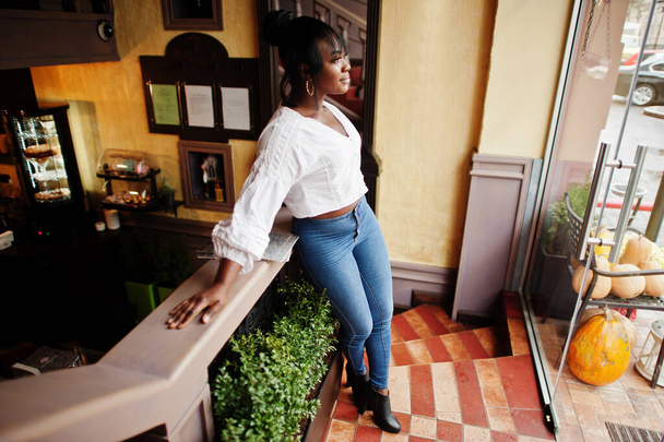 Stylish african american women in white blouse and blue jeans posed at cafe with newspaper. - Foto, Imagem