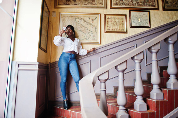 Stylish african american women in white blouse and blue jeans posed at cafe with newspaper. - Foto, afbeelding