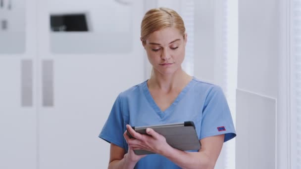 Portrait of smiling female doctor wearing scrubs using digital tablet in hospital corridor - shot in slow motion - Πλάνα, βίντεο