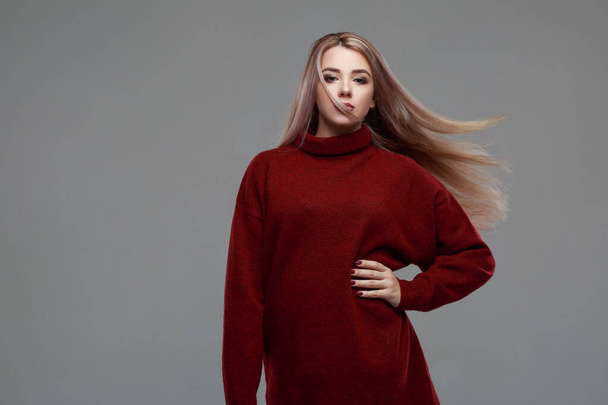 Beautiful blonde girl with long hair flying in the wind in a warm burgundy sweater isolated on a gray background. - Foto, Bild