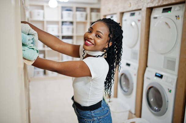 Cheerful african american woman with towels in hands near washing machine in the self-service laundry. - Foto, immagini