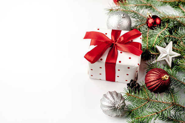Vánoční kompozice na bílém pozadí s bílou dárkovou krabičkou s červenou stuhou s jedlovými větvemi, hračky. Zobrazit top s kopírovacím prostorem pro vaše gratulace - Fotografie, Obrázek