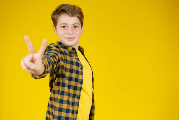 Glücklicher kleiner Junge zeigt Siegesgeste auf gelbem Hintergrund - Foto, Bild
