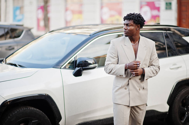 Stylish afro man in beige old school suit against modern hybrid electro suv car. Fashionable young African male in casual jacket on bare torso. - Foto, immagini