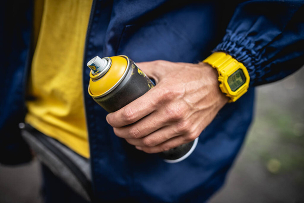 The artist in a blue jacket and a yellow watch holds a spray can of yellow paint in his hand - Foto, Bild