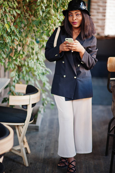 Charming african american woman model in black jacket, hat and waist bag relaxing in cafe during free time with mobile phone. - 写真・画像