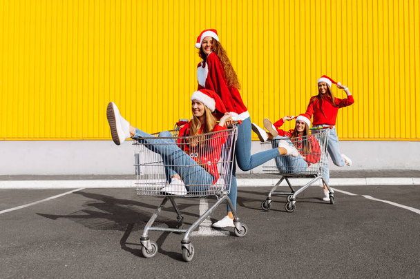 Young happy friends in Christmas sweaters and Santa Claus hats are having fun on a shopping trolley, against the background of a yellow wall of a shopping center, shopping concept, Christmas, New Year - Фото, изображение