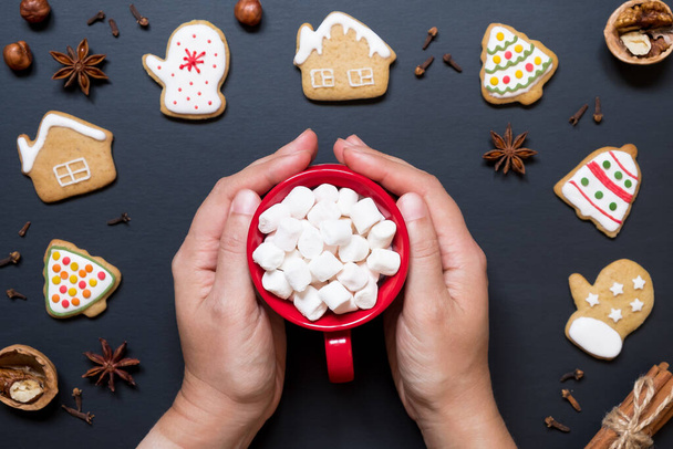 Hands hold red ceramic cup with cocoa and marshmallows on background of christmas cookies. - Foto, Bild
