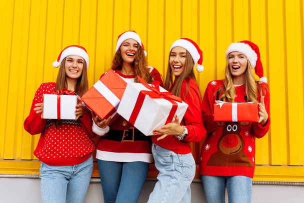 Happy excited group of friends dressed in red sweaters and santa claus hats holding christmas presents while standing near yellow wall near - Foto, Imagem