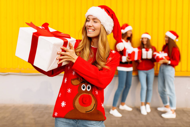 Beautiful smiling young woman wearing santa claus hat with christmas present standing against background of happy friends celebrating christmas outdoors - Fotografie, Obrázek
