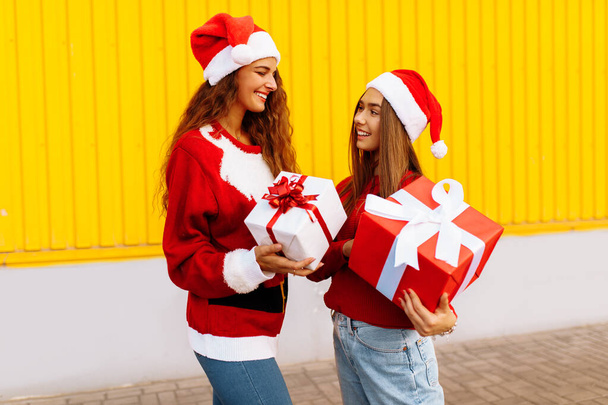 Happy friends in Santa Claus hats, with Christmas gifts, on the background of the yellow wall of the shopping center - Fotografie, Obrázek
