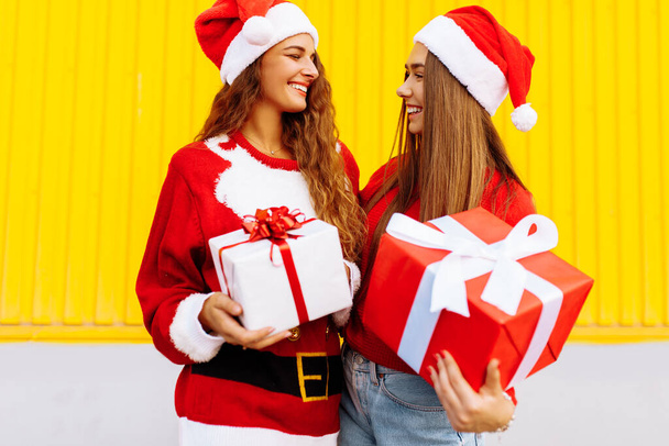 Happy friends in Santa Claus hats, with Christmas gifts, on the background of the yellow wall of the shopping center - Foto, Bild