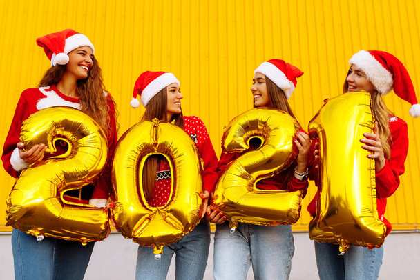 Group of cheerful young women in Santa Claus hats with 2021 balloons on yellow wall background. New Year celebration - Foto, Imagen