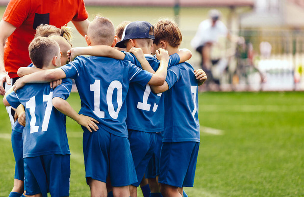 Kids in elementary school sports team with coach. Boys in blue soccer uniforms with white numbers on back. Coach motivate children football players before the game. School sports tournament for yout - 写真・画像