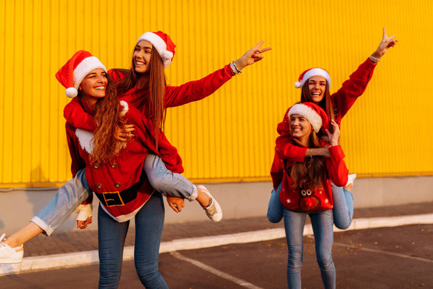 cheerful girlfriends wearing santa claus hats having fun and riding each other outdoors near yellow wall - Zdjęcie, obraz
