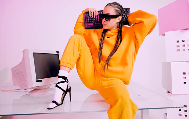 Modern girl in sunglasses sitting with obsolete computer and keyboard on desk in office. Colored Neon light - Фото, изображение