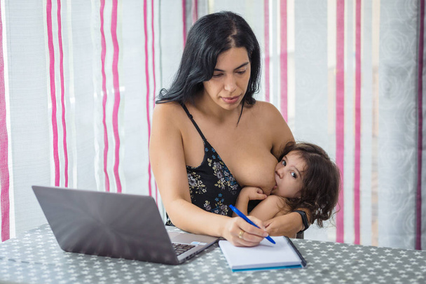 Мать телеграфирует за обеденным столом на ноутбуке, кормя грудью свою девочку, которая сидит у нее на коленях - Фото, изображение
