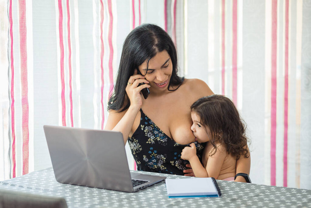 Мать телеграфирует за обеденным столом на ноутбуке, кормя грудью свою девочку, которая сидит у нее на коленях - Фото, изображение