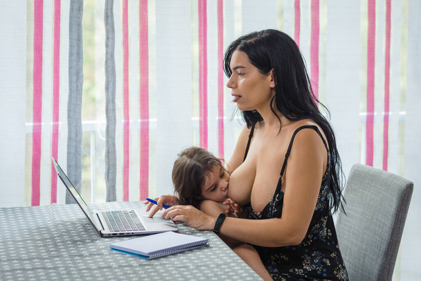 Mutter arbeitet am Esstisch auf einem Laptop, während sie ihr kleines Mädchen stillt, das auf ihrem Schoß sitzt - Foto, Bild