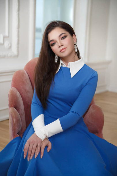 Beautiful playful brunette girl in a blue dress sits in a light interior. - Fotoğraf, Görsel
