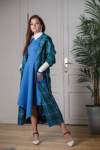 Beautiful brunette girl in a blue dress and a stylish trench coat in a light interior with a palm tree. - Zdjęcie, obraz