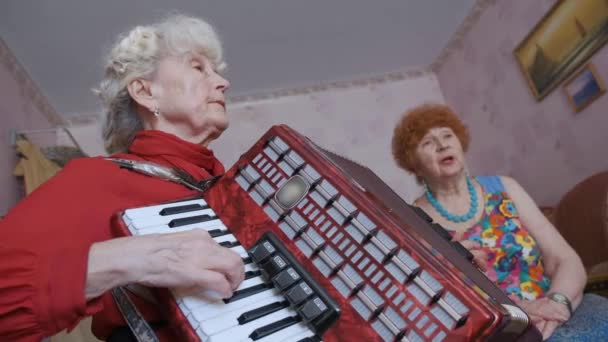 Senior mujer juega acordeón mientras amigo canta en la habitación - Imágenes, Vídeo
