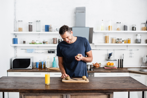 Glimlachende man kijkt naar kiwi op snijplank bij sinaasappelsap in glas op tafel in de keuken  - Foto, afbeelding