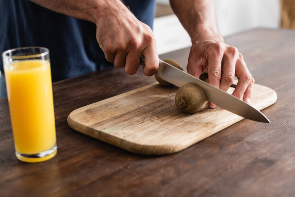 Cropped view of man cutting kiwi near glass of orange juice on blurred foreground  - Foto, afbeelding