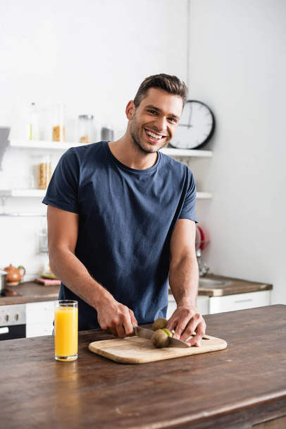 Smiling man looking at camera while cutting kiwi on cutting board near orange juice in kitchen  - Photo, image