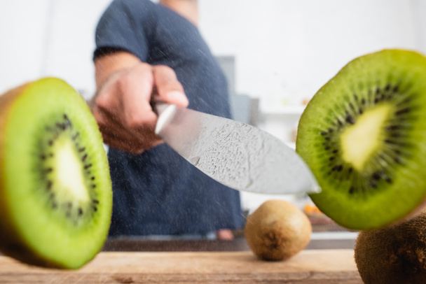 Close up view of halves of kiwi near knife in hand of man on blurred background  - Fotografie, Obrázek