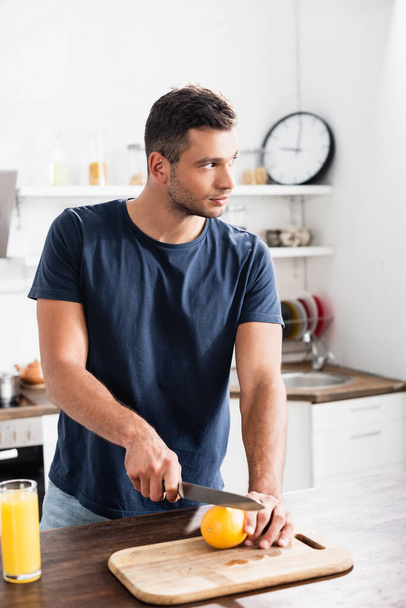 Young man looking away while cutting orange near glass of orange juice on blurred foreground  - Foto, Imagen
