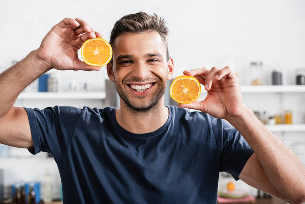 Cheerful man holding halves of orange and looking at camera in kitchen  - 写真・画像