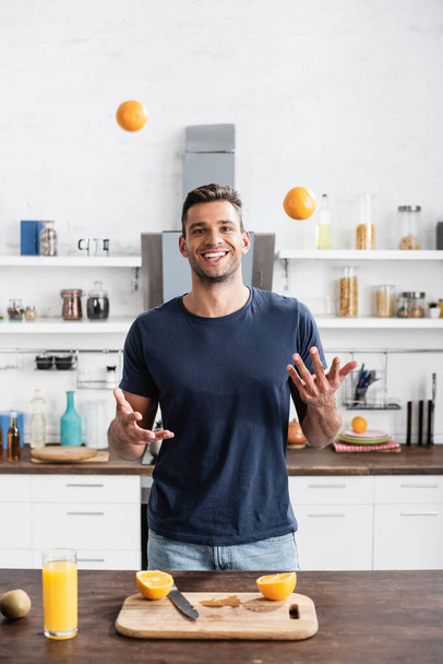 Cheerful man throwing oranges near cutting board and glass of orange juice  - 写真・画像