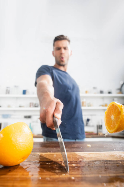 Close up view of halves of orange on wet cutting board and knife in hand of young man on blurred background  - Photo, Image