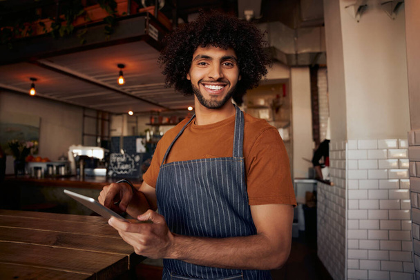 Portrait of handsome young male waiter standing in cafe wearing apron holding digital tablet - Foto, Imagem