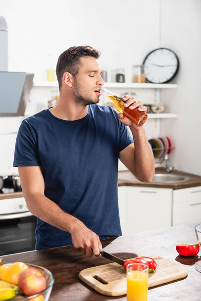 Man drinking beer while cutting bell pepper near orange juice and fruits on blurred foreground  - Foto, Imagen