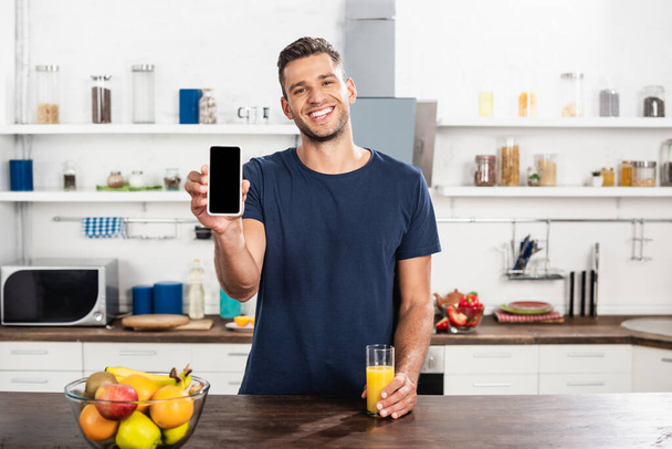 Homem sorridente mostrando smartphone com tela em branco perto de vidro de suco de laranja e frutas na cozinha  - Foto, Imagem