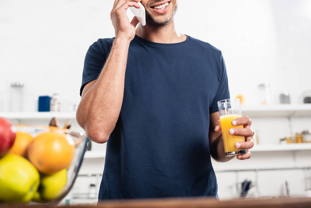 Cropped view of smiling man with glass of orange juice talking on smartphone near fruits on blurred foreground in kitchen  - Фото, изображение