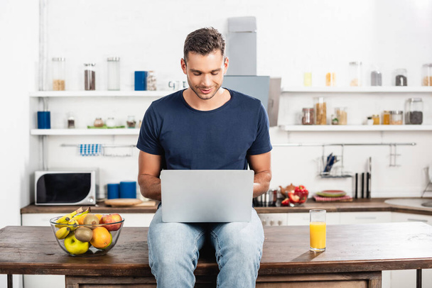 Young man using laptop near glass of orange juice and fresh fruits in kitchen  - Foto, Imagen