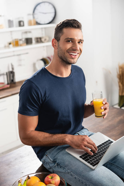 Mosolygó ember egy pohár narancslé laptoppal, miközben ül gyümölcsök közelében konyhaasztalon  - Fotó, kép