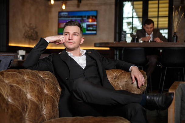 A young elegant man in a dark classic three-piece suit sits in a leather armchair in a rest area of a restaurant. - Foto, Imagem